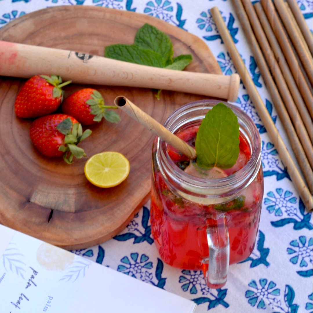 Mason jar with red drink, strawberries, and mint leaves on a table with wooden straws and a plate of strawberries and lemon.