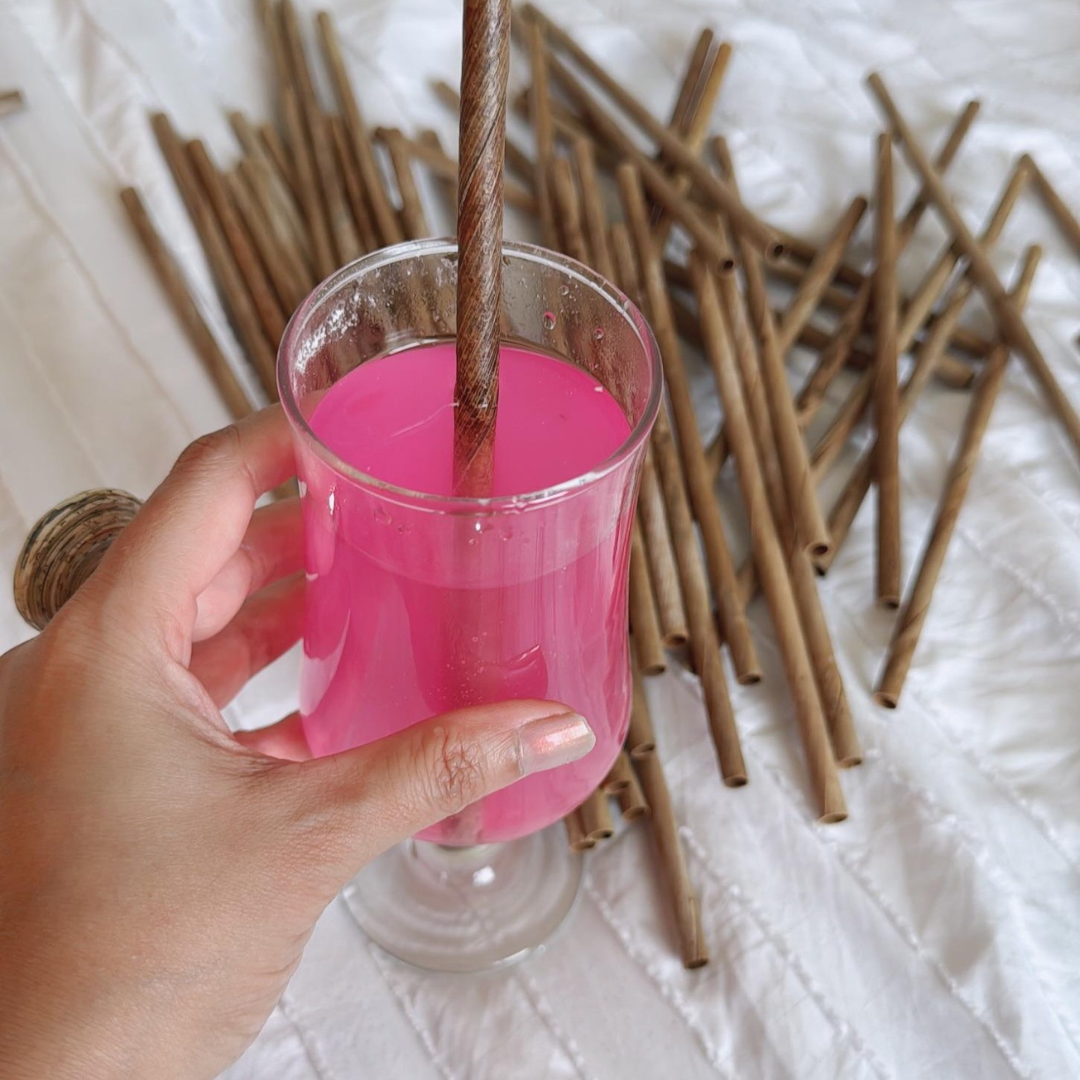 Hand holding a glass of pink liquid with a straw, surrounded by wooden straws on a white surface.